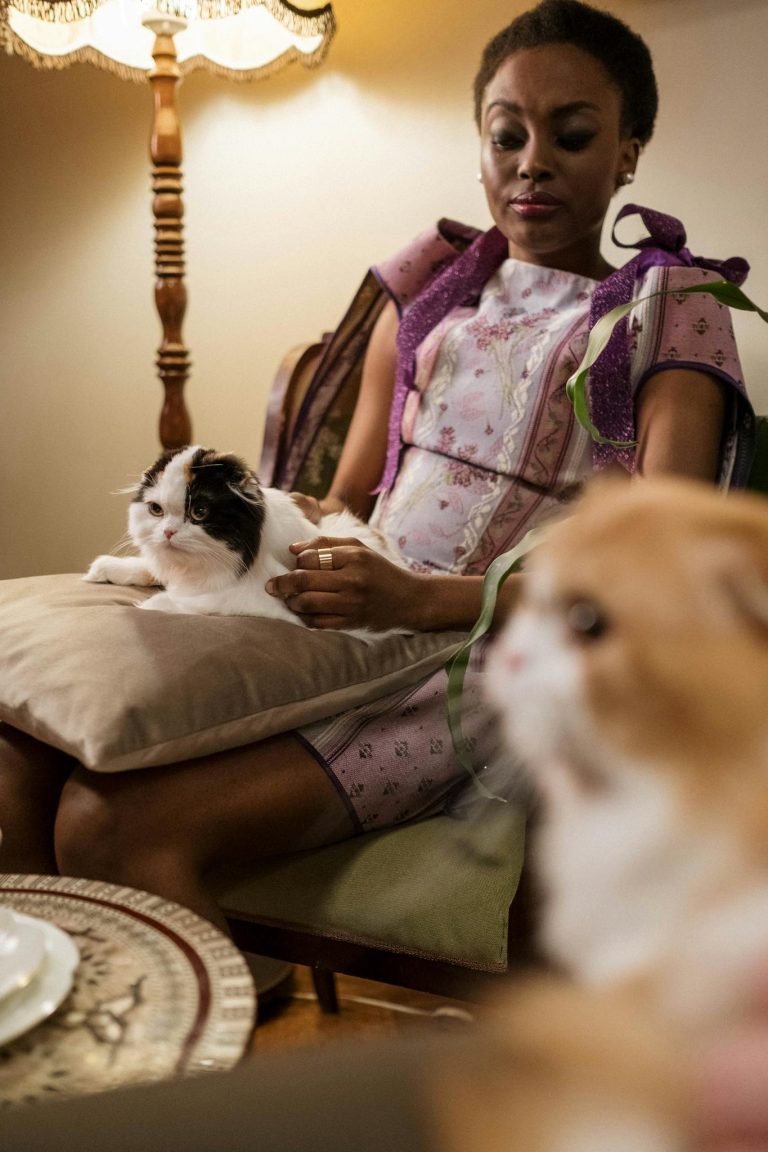African American woman in patterned dress sitting with cats indoors, cozy lamp-lit setting.